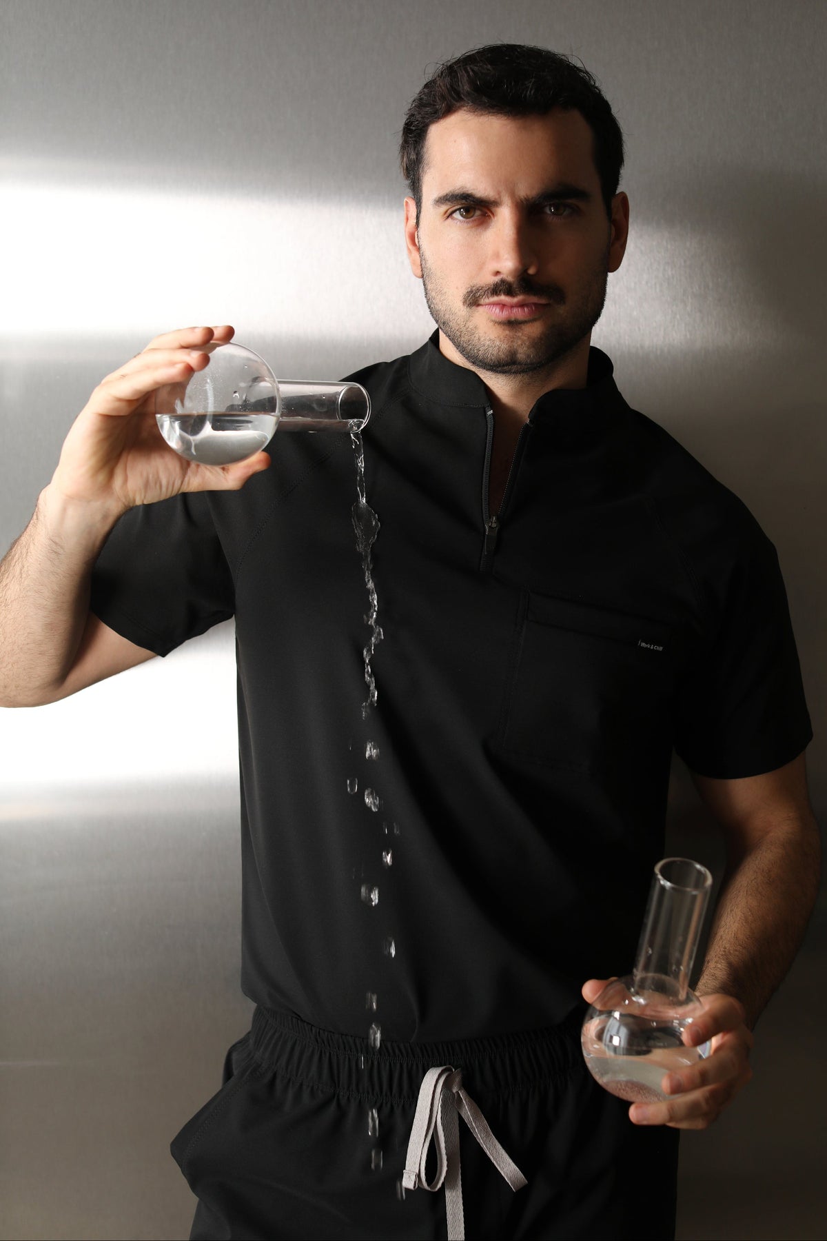 Man wearing the Harry Black W&C Shield™ scrub top while pouring clear liquid from a laboratory flask.