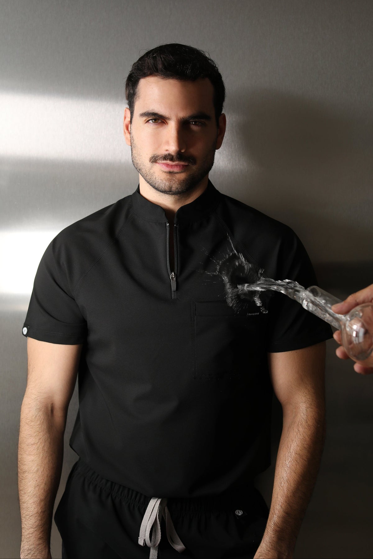 Man wearing the Harry Black W&C Shield™ scrub top during a water-repellency demonstration with clear liquid splashing across the chest.